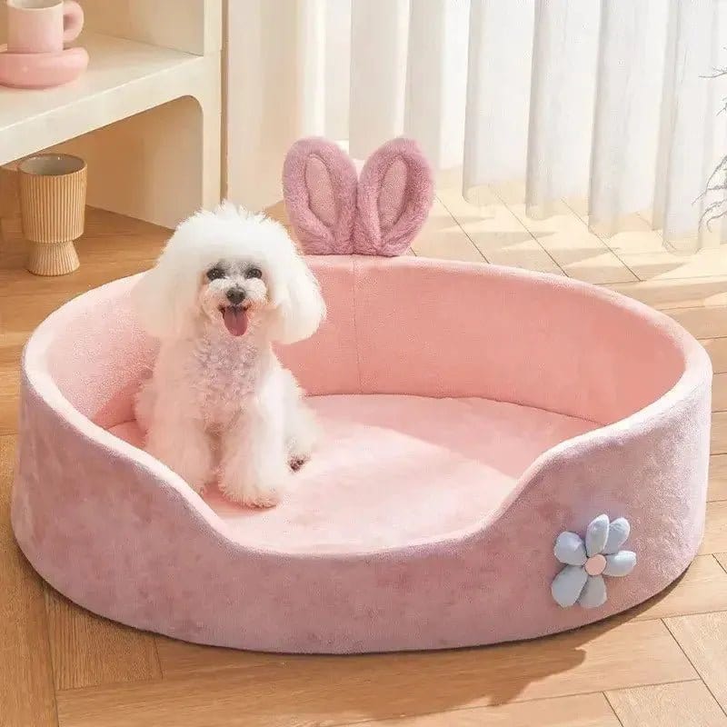 A white dog sitting on a pink washable dog kennel bed with a decorative element resembling rabbit ears on the top corner and a blue flower on the side.